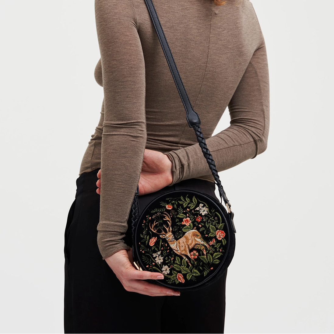 Person holding a round embroidered bag with floral and animal designs on a plain background