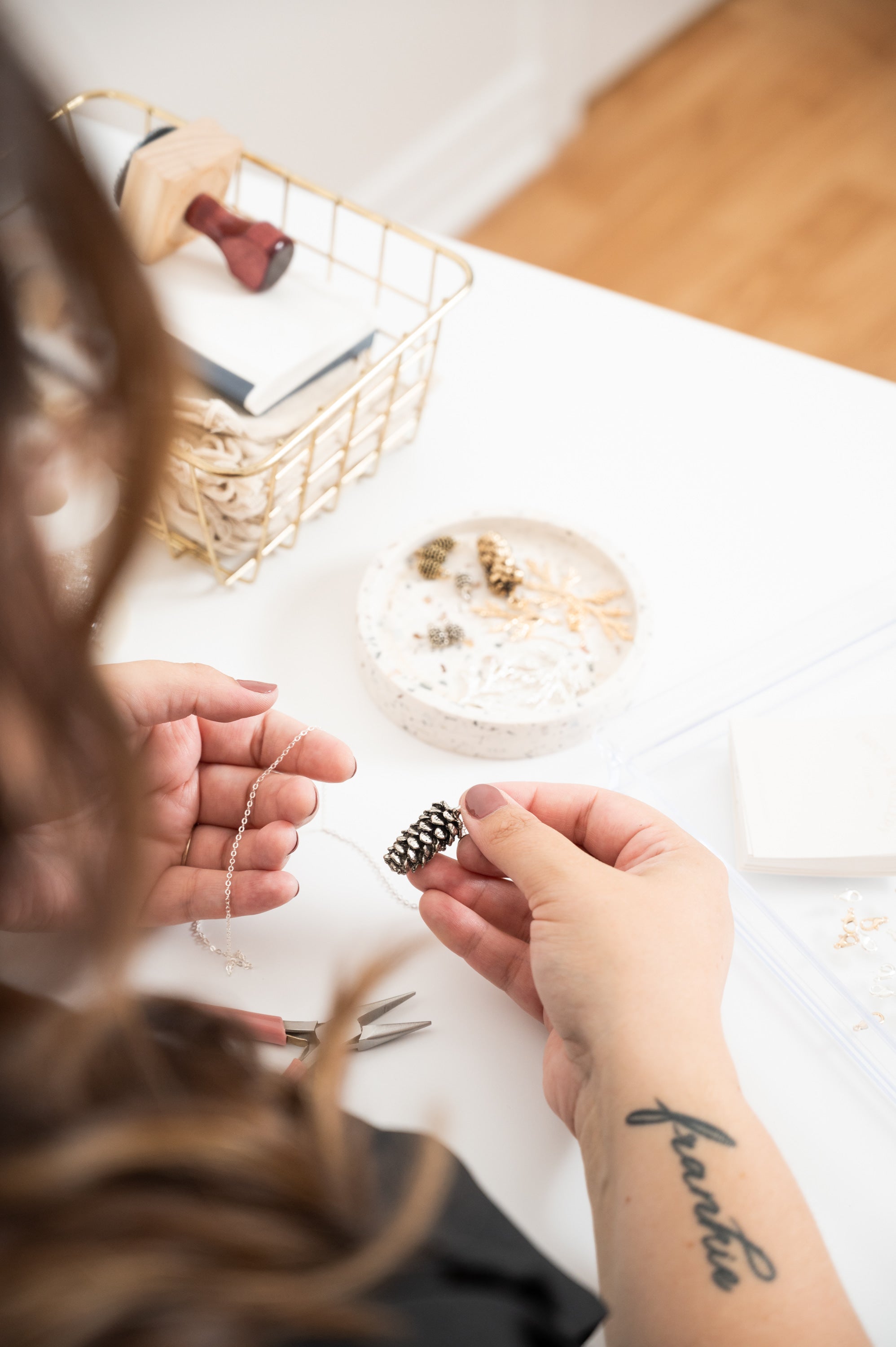 A person working with jewelry on a table with a basket and container in the background.