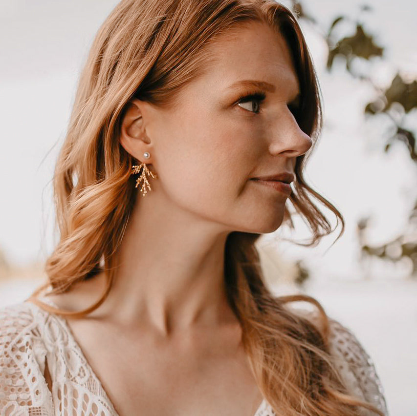A red-headed bride with her hair curled and tucked behind her ear looking off to the right. She is wearing a gold juniper branch and pearl ear jacket.